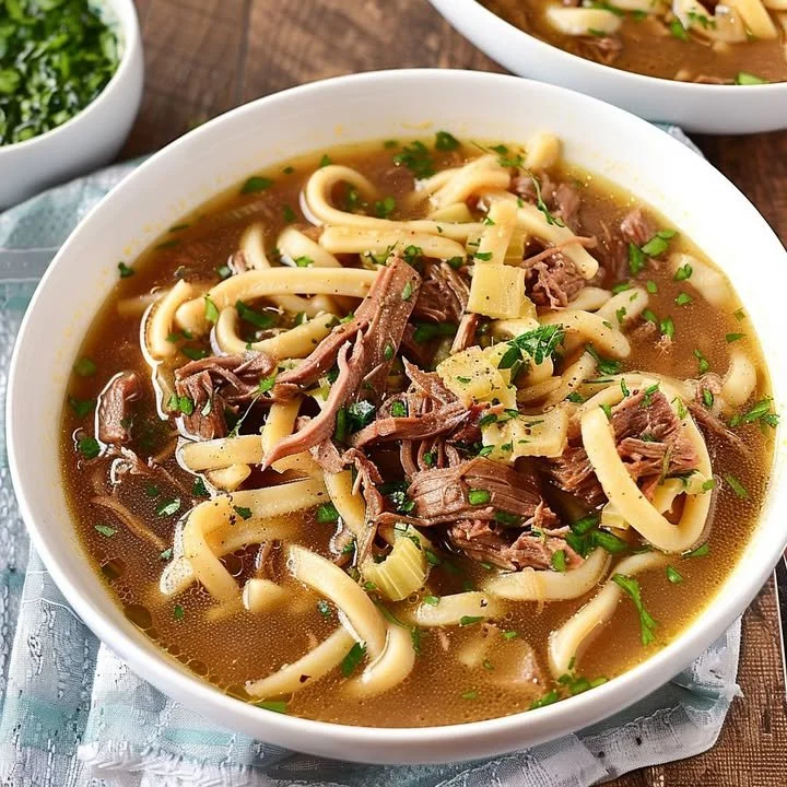Bowl of slow-cooked beef noodle soup with fresh herbs and vegetables.